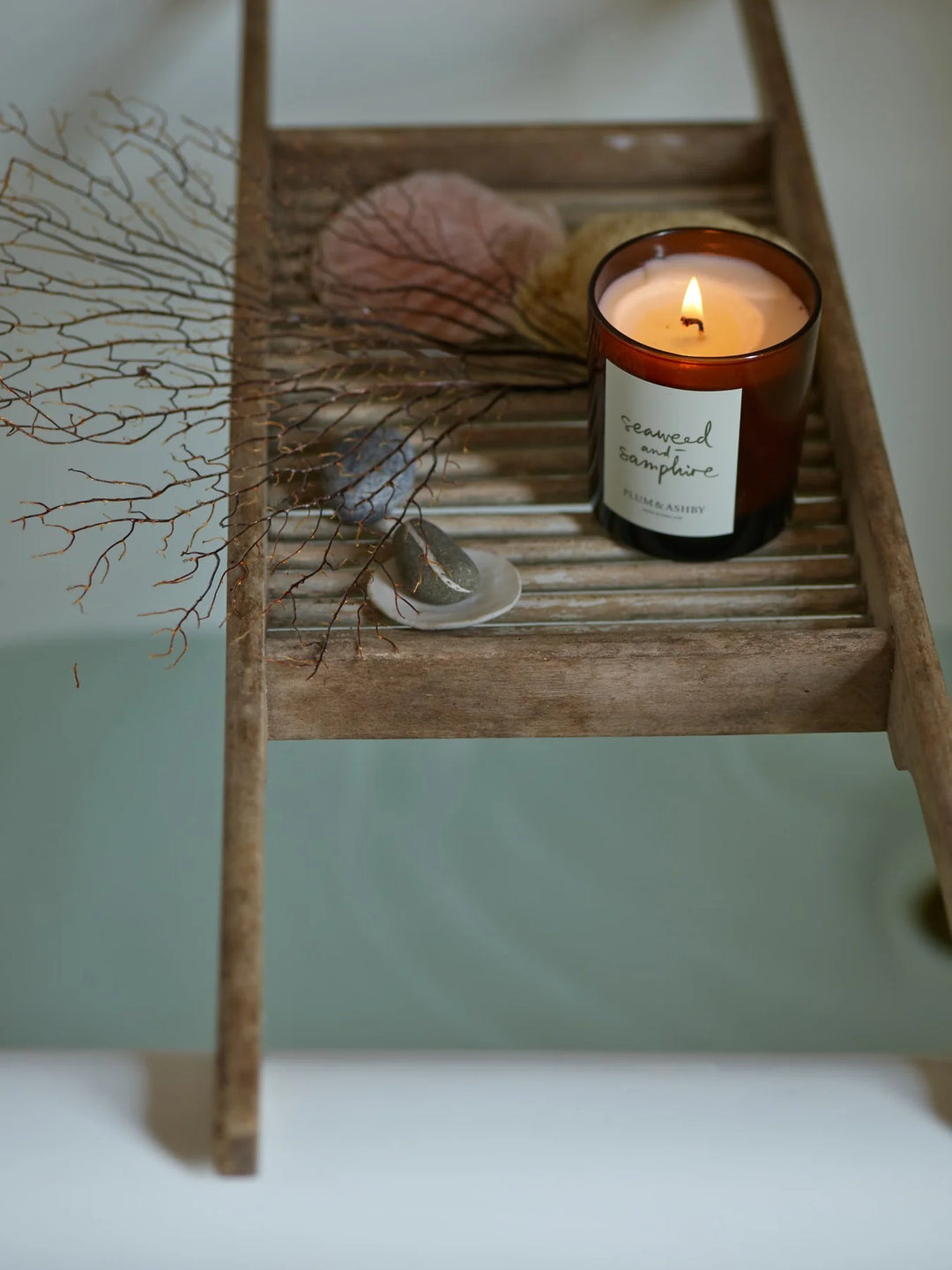 Lit seaweed and samphire candle on a timber rack over a bathtub with coral and stones
