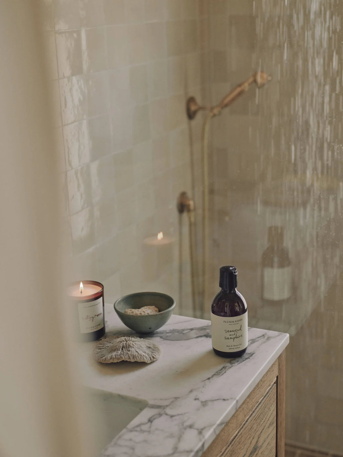 Amber bottle of seaweed and samphire shower gel on a marble bathroom bench with a lit candle and shower in the background