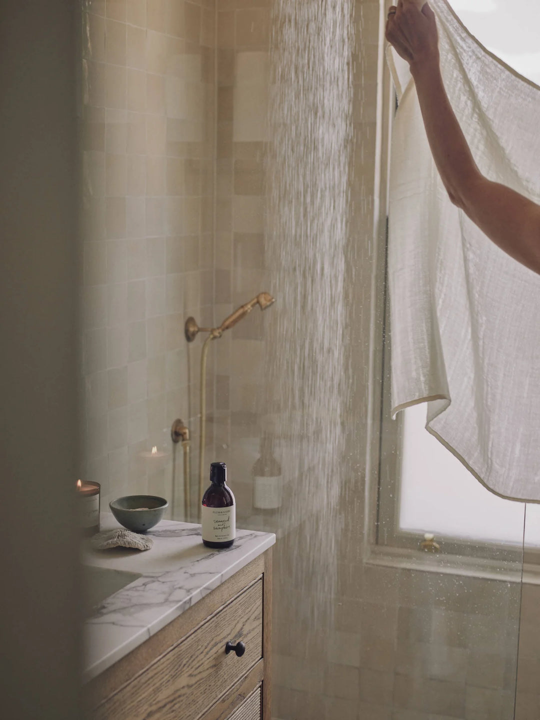 Amber bottle of seaweed and samphire shower gel on a marble bathroom bench with a lit candle and shower in the background