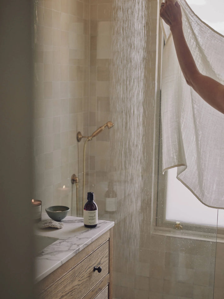 Amber bottle of seaweed and samphire shower gel on a marble bathroom bench with a lit candle and shower in the background