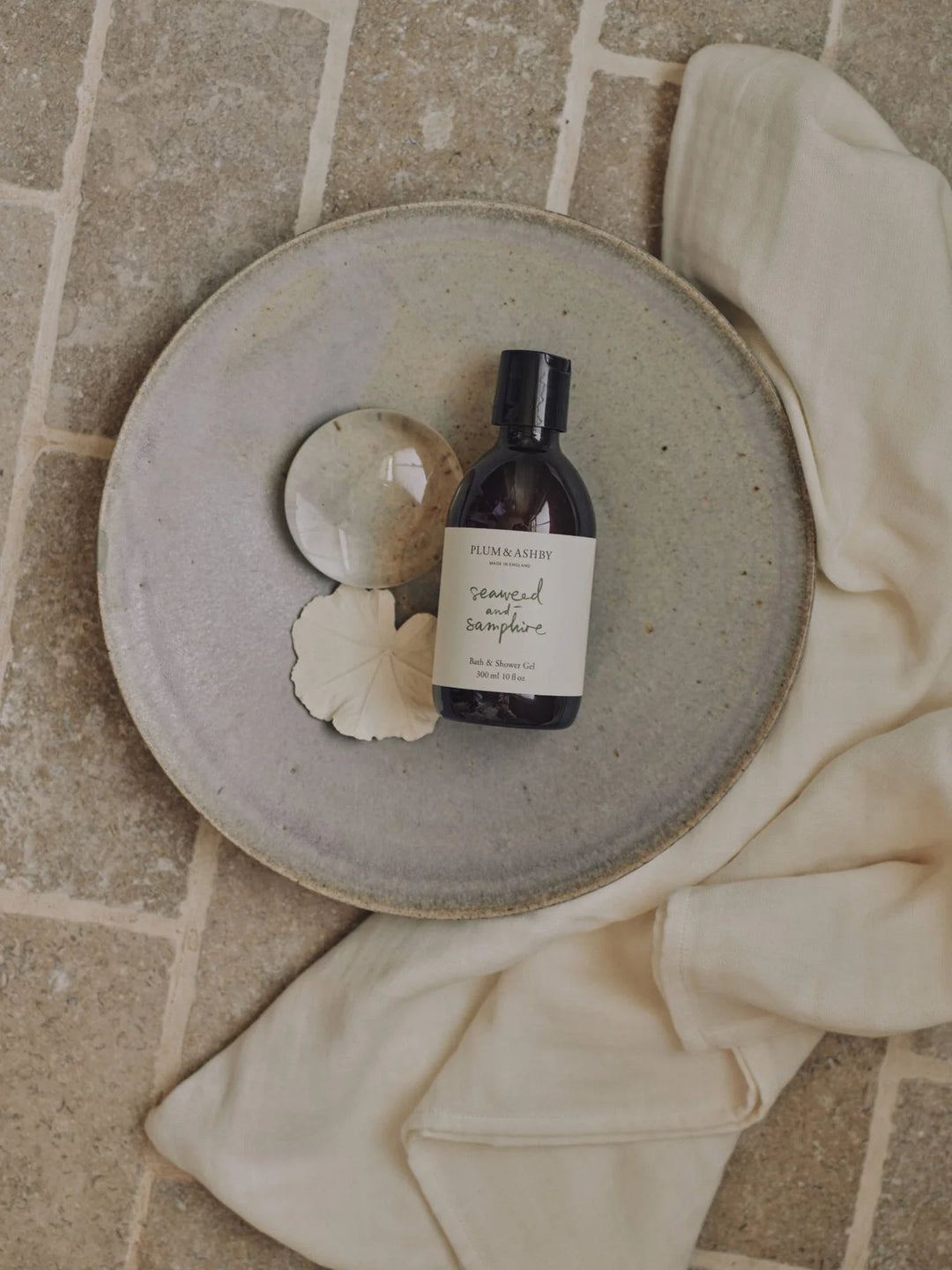 Amber bottle of seaweed and samphire shower gel on a stone plate with cream linen on a brick surface