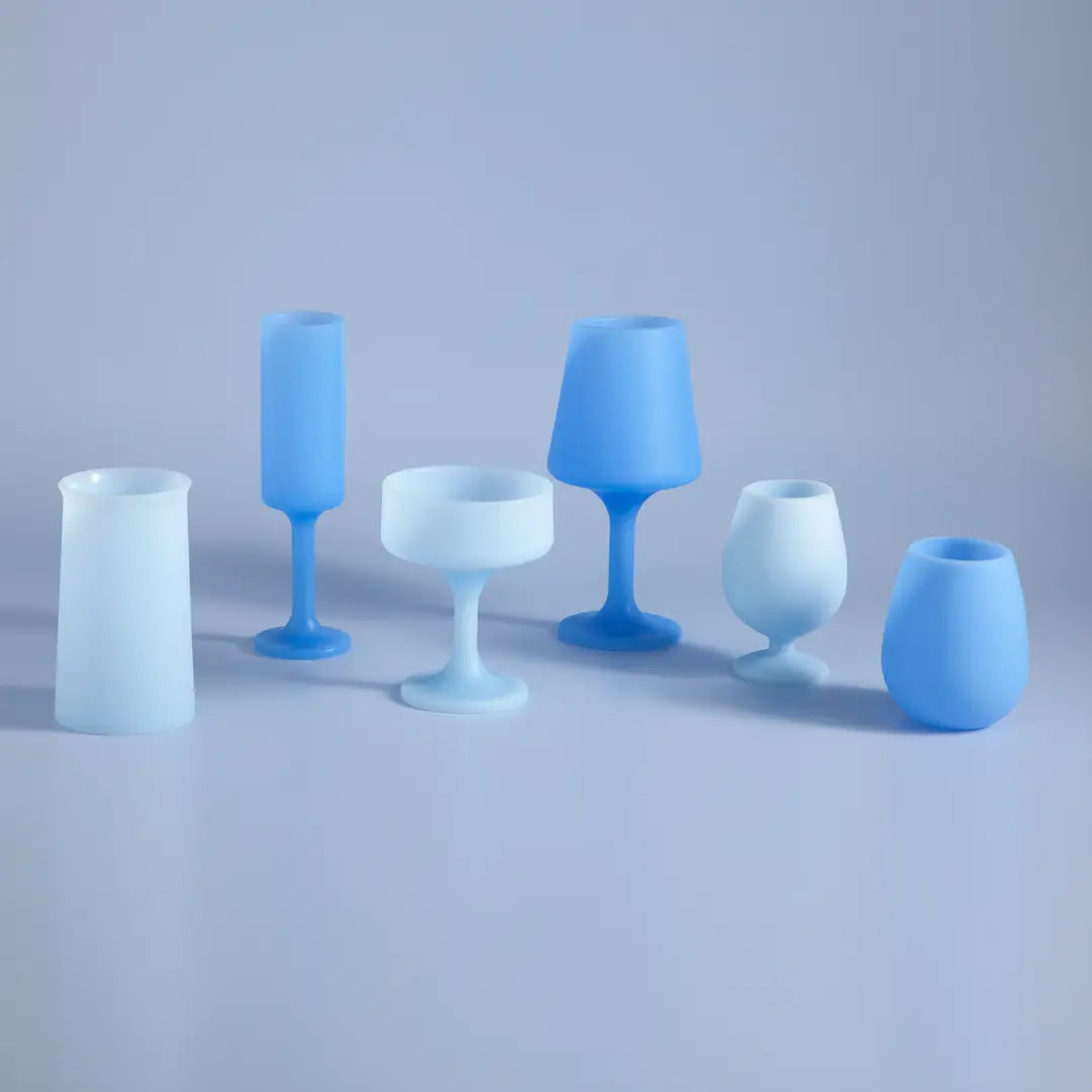 Six matte blue and white silicone glassware pieces, including tumblers, goblets, and a coupe glass, arranged on a light gray studio surface.