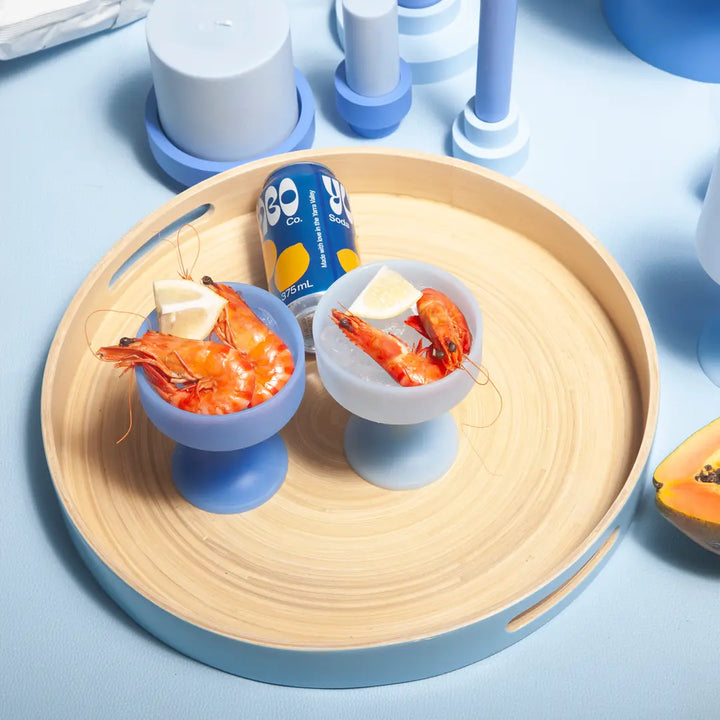 Two small blue silicone cocktail glasses with prawns and lemon wedges on a wooden tray with a can of soda.