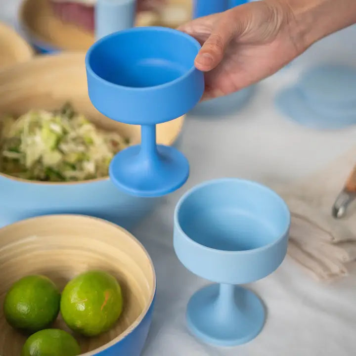 A hand holds a bright blue silicone cocktail glass above a matching light blue pedestal bowl, with more blue bowls and limes in a softly blurred kitchen setting.