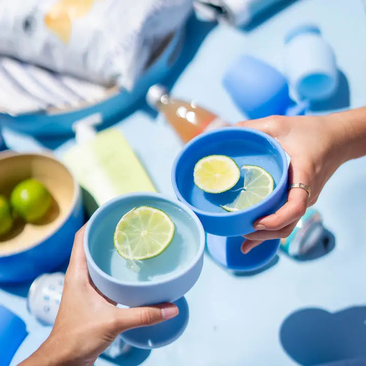 Two hands holding blue silicone cocktail coupe glasses filled with water and lime slices, clinking together on a light blue surface with a blurred outdoor picnic-style background.