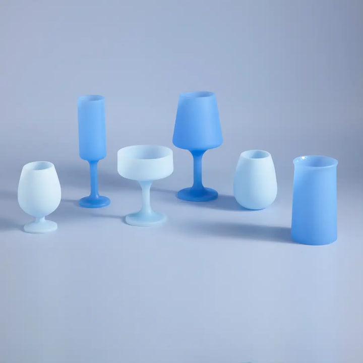 Six matte blue and white glass-like drinking vessels in different shapes, arranged on a smooth light gray studio surface.