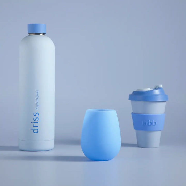 Three drinkware products arranged on a light gray studio surface: a tall matte white insulated bottle with a blue cap and blue branding, a small translucent light blue tumbler, and a reusable cup with a blue lid and blue sleeve.