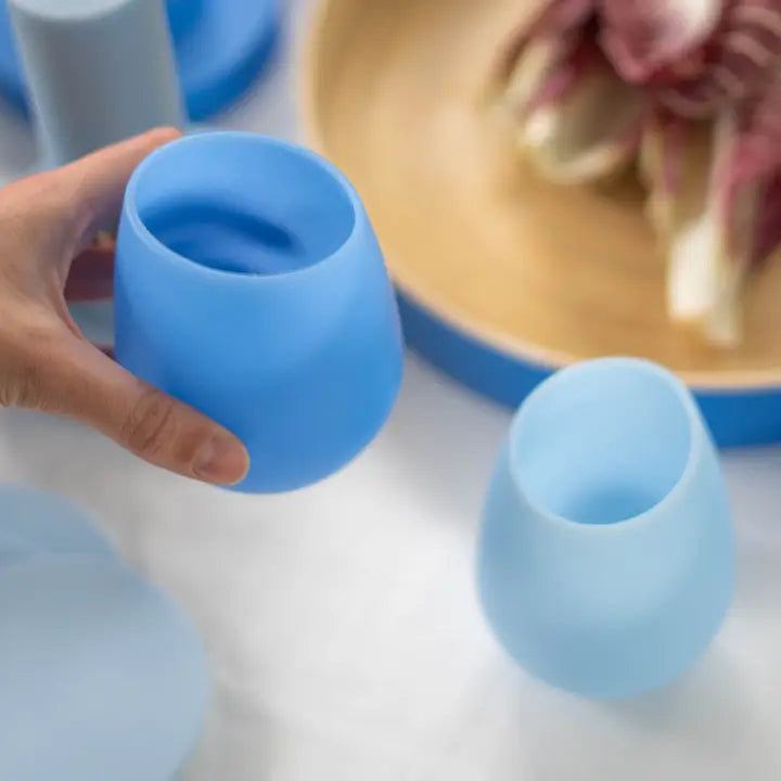 A hand holds a light blue silicone stemless glass, with another matching glass and a light blue lid on a white surface in the foreground, and a wooden plate in the blurred background.