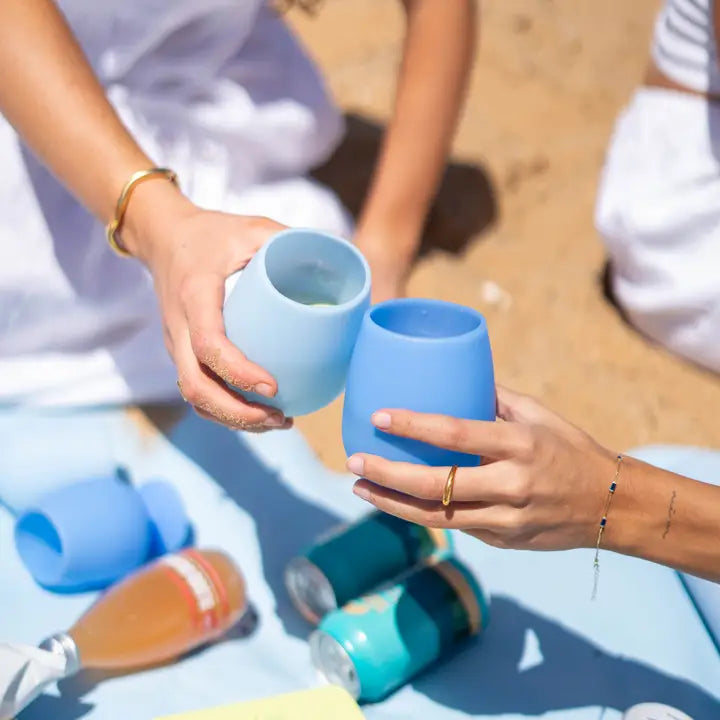 Two people clinking reusable silicone cups at a beach picnic. One cup is light blue and the other is medium blue, with other blue cups and cans on a blanket in the sandy background.