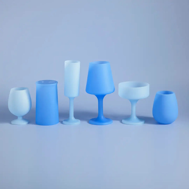 Six matte blue and light blue silicone drinking glasses arranged in a row against a soft blue-gray studio background.