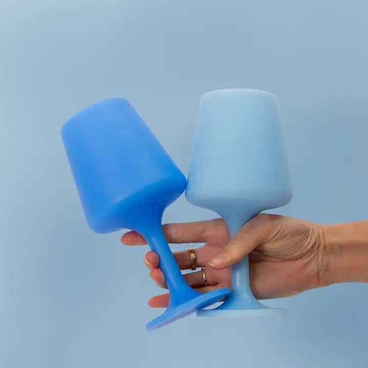 Two silicone wine glasses in shades of blue, one held by a hand and one standing upright, against a solid light blue background.