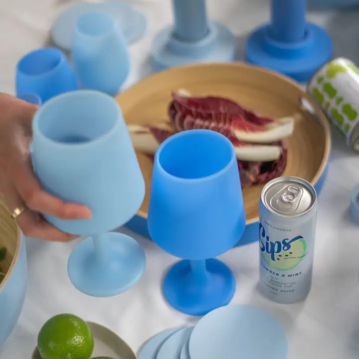 A set of silicone wine glasses in light blue and medium blue, shown on a white tablecloth with food props and a can of Sips water.