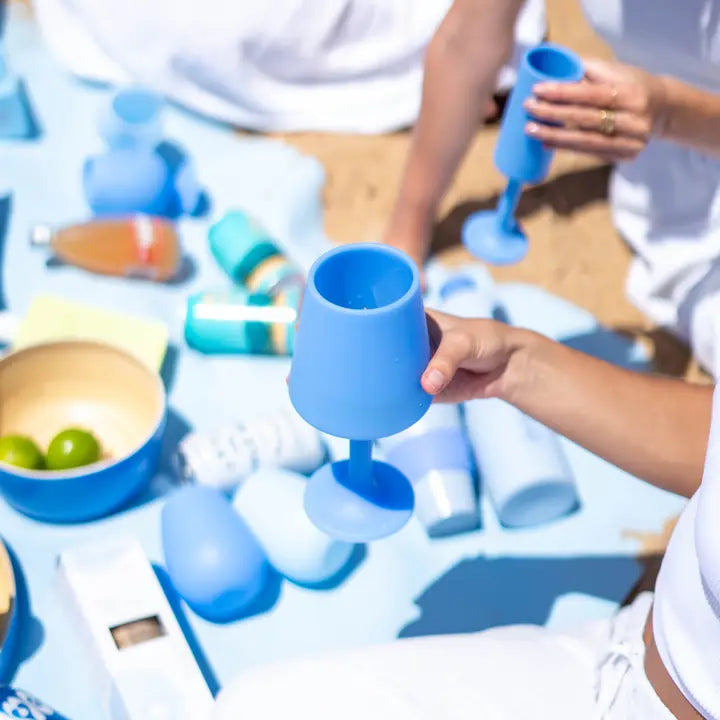 A person outdoors holds a bright blue silicone drinking glass with a stem and wide base, surrounded by similar blue vessels and bottles on a light blue blanket.