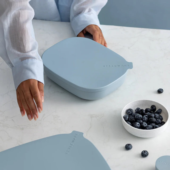 A light blue silicone lunch box with a matching lid is shown on a white marble tabletop, with a person&