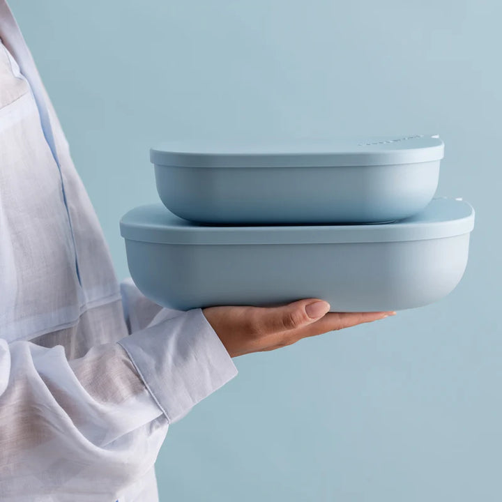 A person in a white shirt holds two stacked light blue oval containers with lids against a solid light blue background.