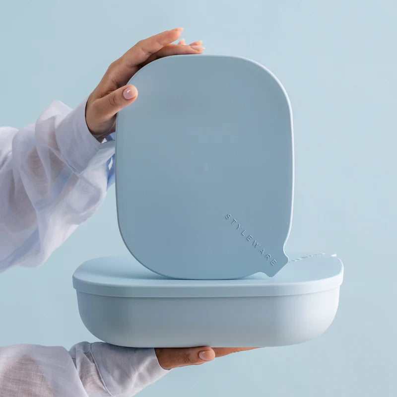 A person in a white blouse holds a light blue silicone mouthguard or dental dam over a matching light blue storage container, with the brand name &
