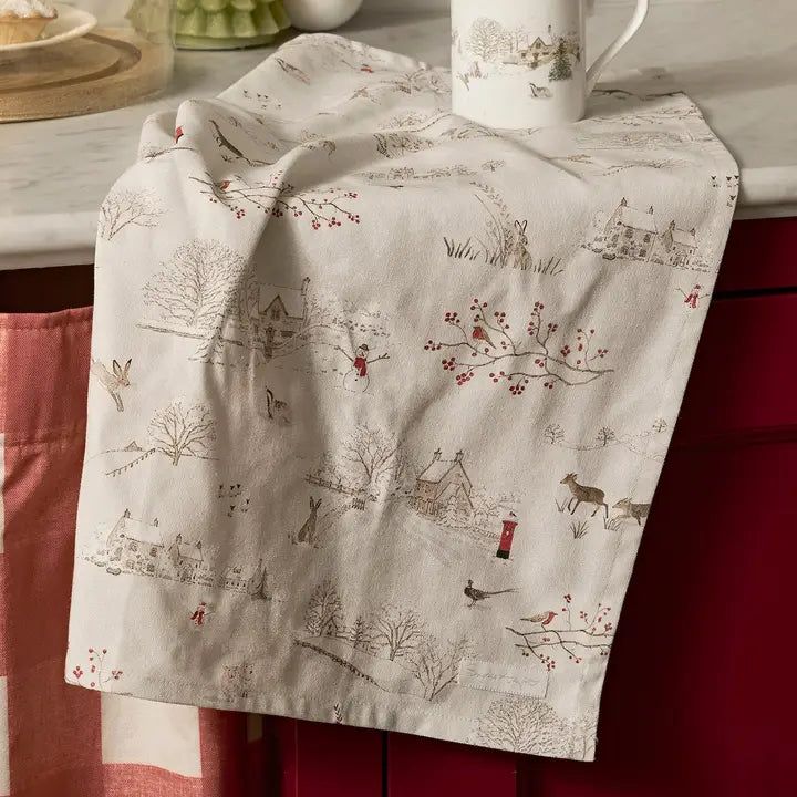Cream coloured Christmas themed tea towel with winter images including red robin and branch with berries, snowy roofed houses, snowman, reindeer, hare and more. Draped on a kitchen benchtop.