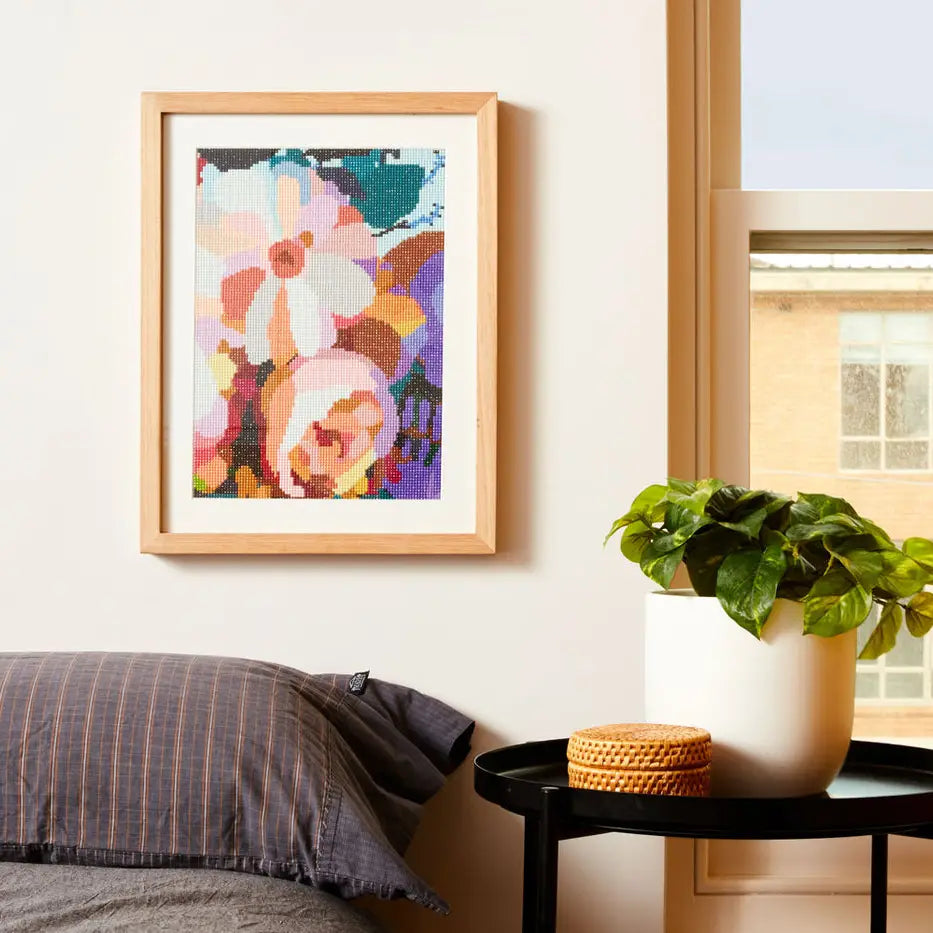 A framed floral sparkle artwork with a light wood frame hangs on a beige wall in a bedroom setting, next to a window, a potted green plant on a black side table, and a bed with gray striped pillows.