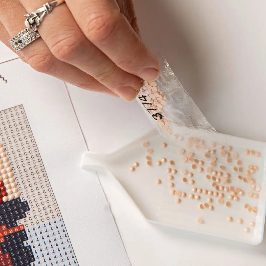 A close-up of a hand pouring small beige rhinestones from a clear plastic bag into a white tray, with a diamond painting pattern sheet partially visible on the left.