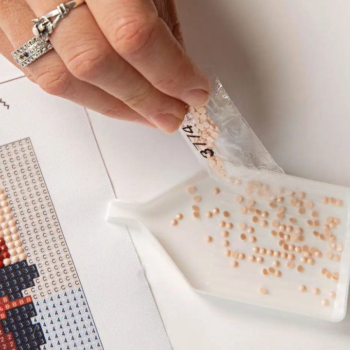 A close-up of a hand pouring small beige rhinestones from a clear plastic bag into a white tray, with a diamond painting pattern sheet partially visible on the left.