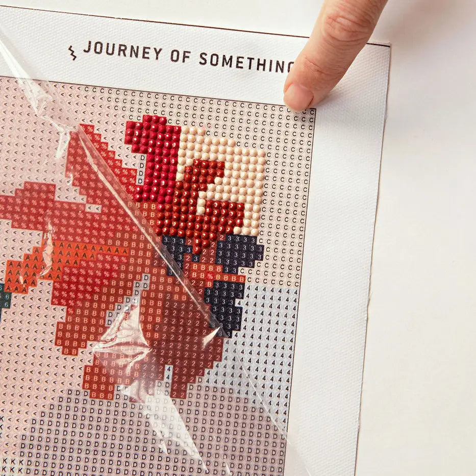 A close-up of a sparkle art canvas with a partially completed red, orange, and black design, covered by a clear plastic protective sleeve. A finger is pressing down on the top right corner of the canvas, which has the text &