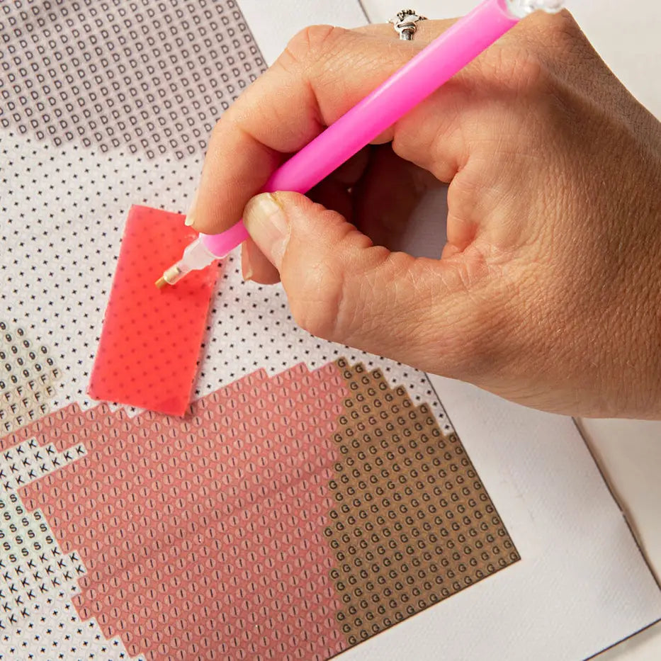 A close-up of a hand holding a pink diamond painting pen and applying it to a red patterned section of a diamond painting canvas.