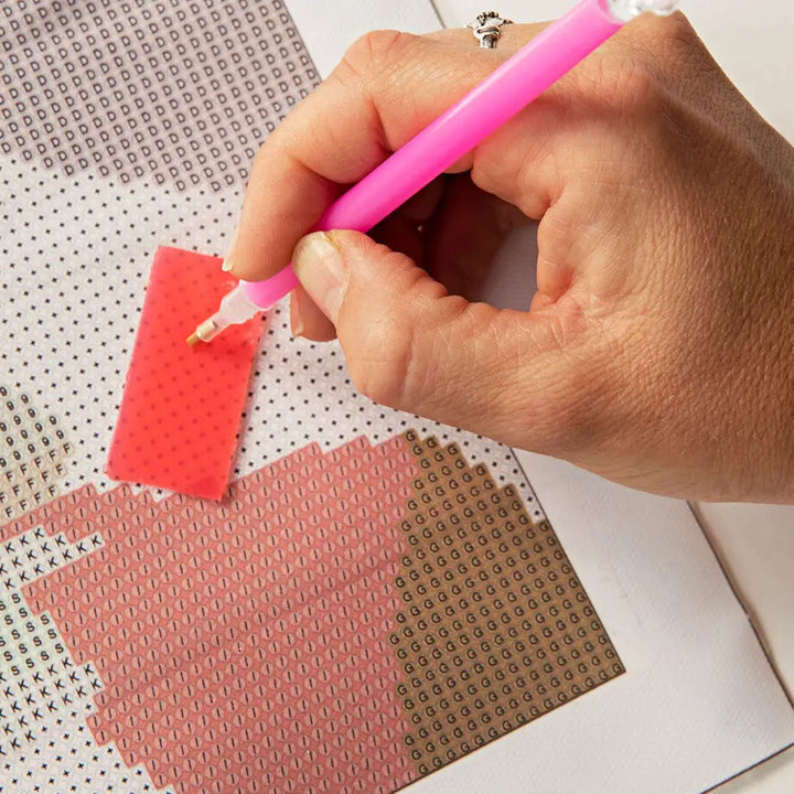 A close-up of a hand holding a pink diamond painting pen and applying it to a red patterned section of a diamond painting canvas.