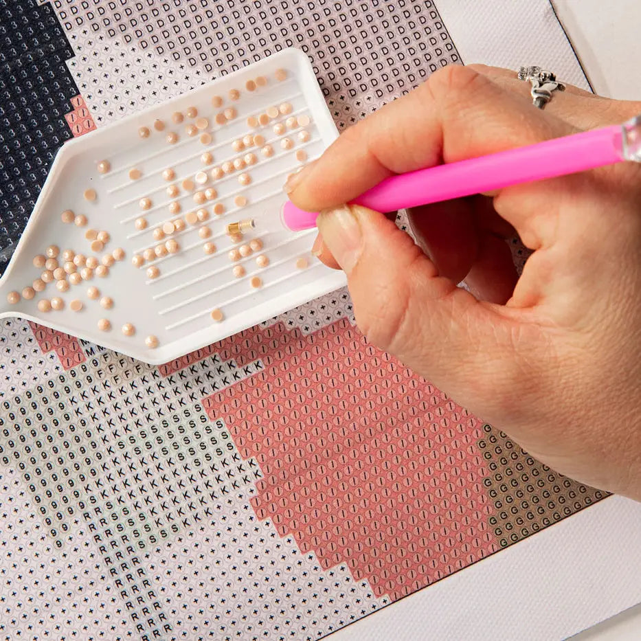 A close-up of a hand holding a pink diamond painting pen over a colorful patterned canvas, with small beige beads arranged in rows on a white tray.