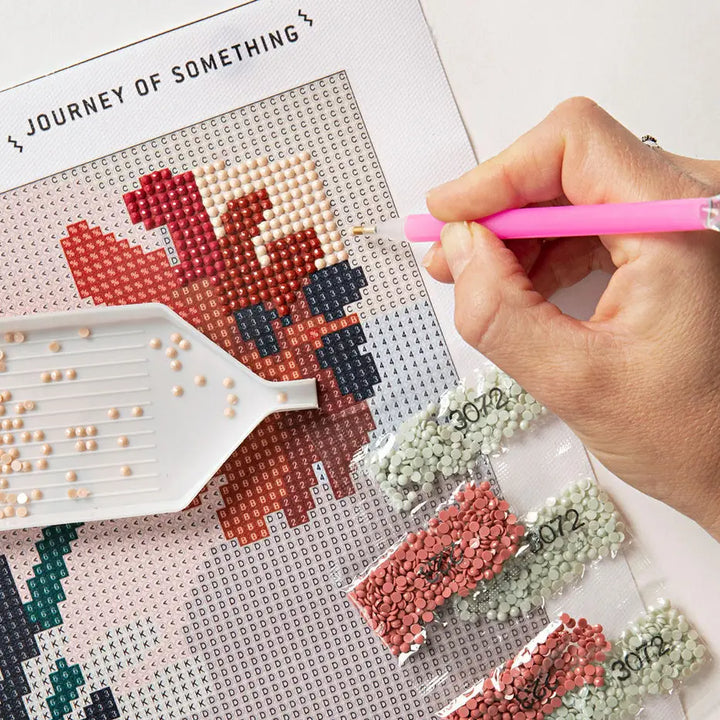 A close-up of a hand using a pink diamond painting pen to apply adhesive beads onto a canvas. The canvas shows a partially completed pixelated design in red, beige, and dark blue.