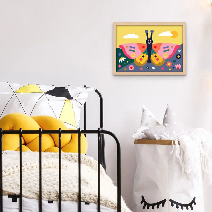 A colorful framed butterfly wall art print hangs on a white wall above a bed with yellow pillows and bedding, and a white storage basket with black eyelash details beside it.