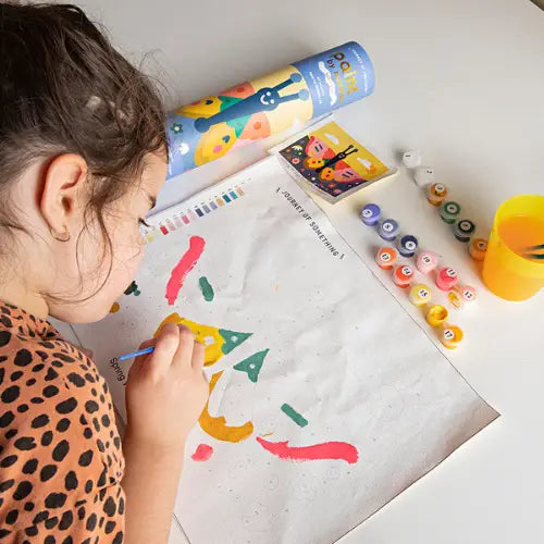 A child painting on a white sheet of paper with a paintbrush, surrounded by colorful paint tubes, a paint palette, and a paint cup on a white table. A blue cylindrical box labeled &