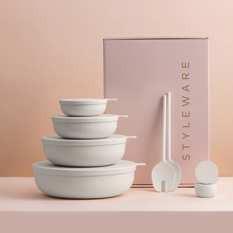 A set of matte whiteplastic bowls with lids, salad servers & mini containers arranged in front of a pink product box labeled STYLEWARE on a soft blush background.
