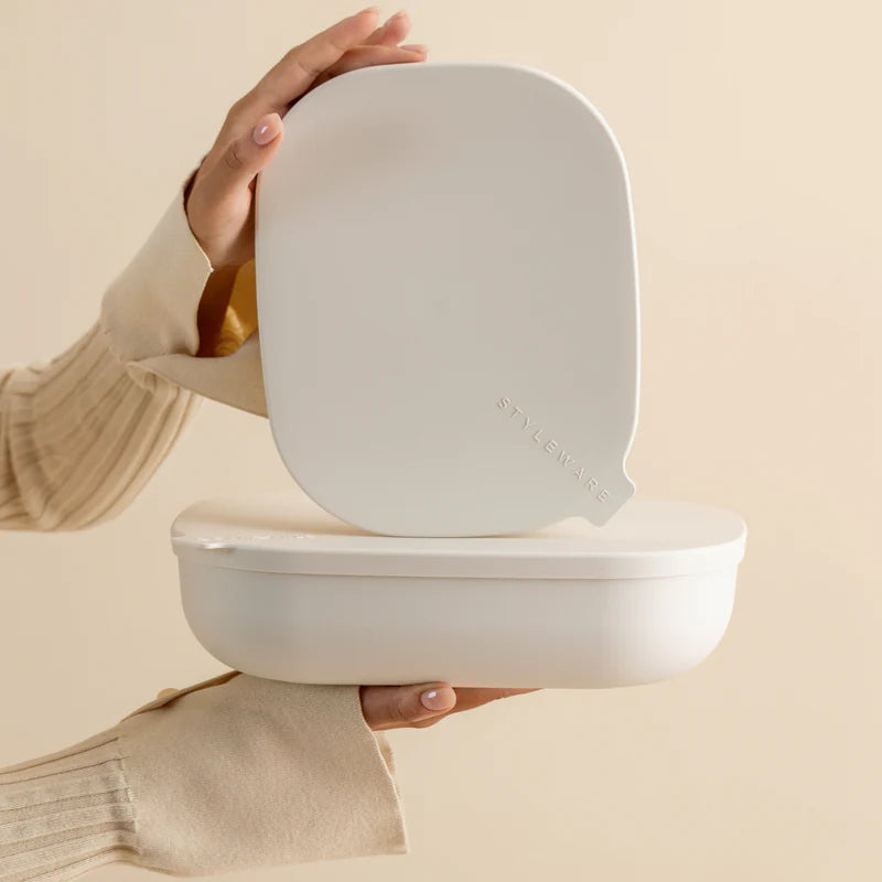 A person in a beige ribbed sweater holds a white oval-shaped container with a matching lid, shown against a soft beige background.