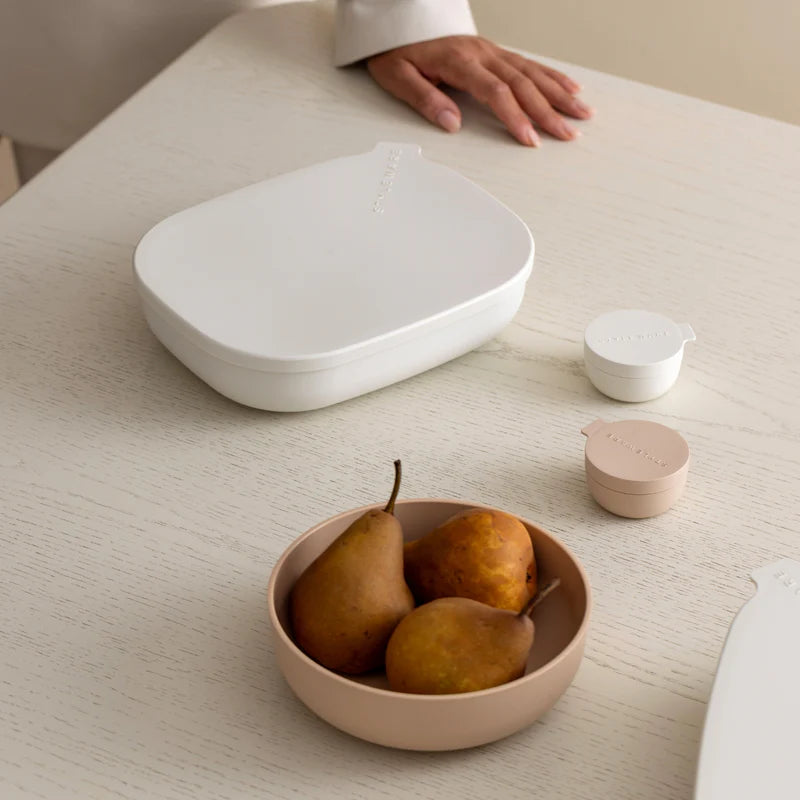 A minimalist white food storage container with a matching lid sits on a light wood table next to a small blush pink bowl containing pears. A hand rests on the table near the lid, and two small round containers are visible.