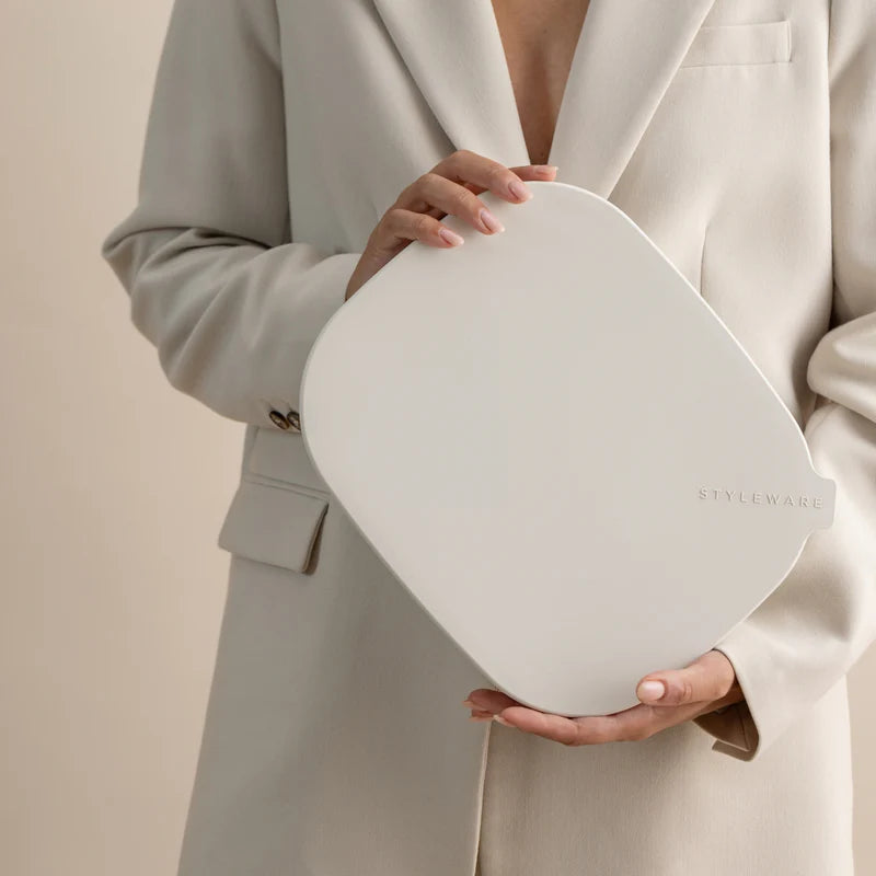 A person in a light beige blazer holds a large, rounded-edged white cosmetic pouch or compact with the brand name &