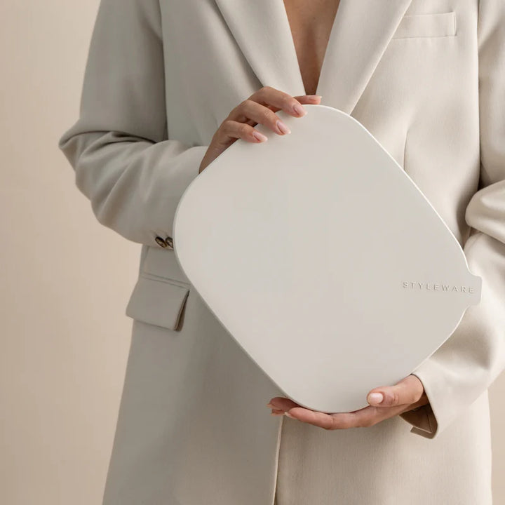 A person in a light beige blazer holds a large, rounded-edged white cosmetic pouch or compact with the brand name &