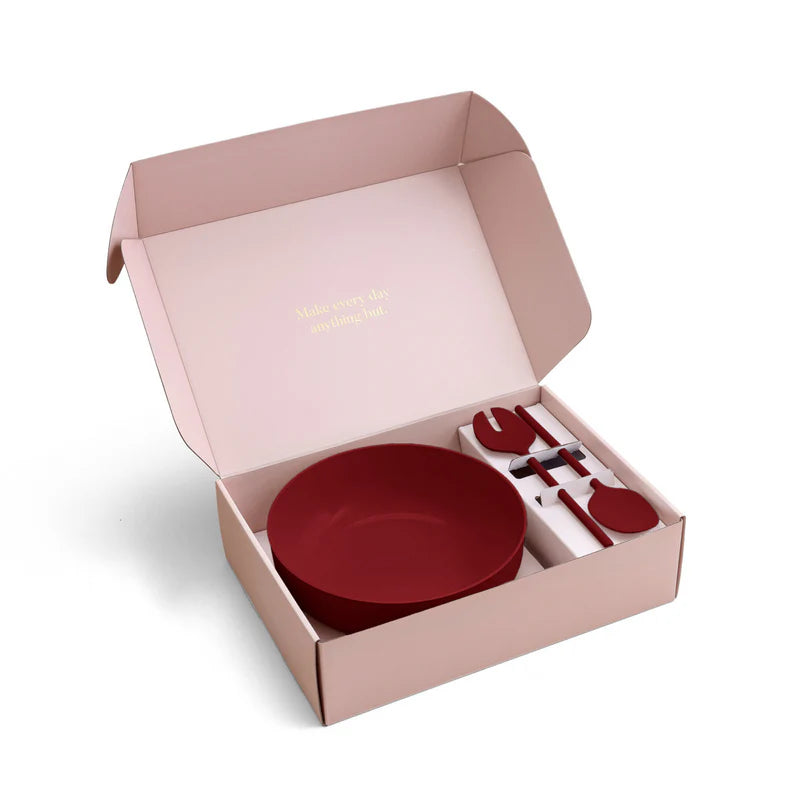 An open blush pink gift box containing a dark red bowl and matching red cutlery on a white background.