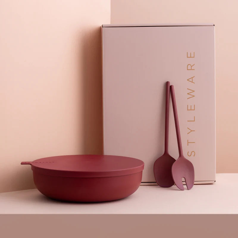 A matte dark red serving bowl with a lid sits on a light beige surface, next to a tall pale pink box labeled STYLEWARE and two matching utensils.