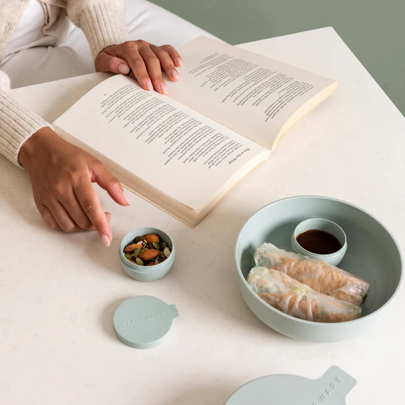 A light sage plastic bento lunch box set on a white table, shown with a person's hands holding an open book. The set includes a main compartment with spring rolls, a small dipping sauce cup, a small snack container, and a lid.