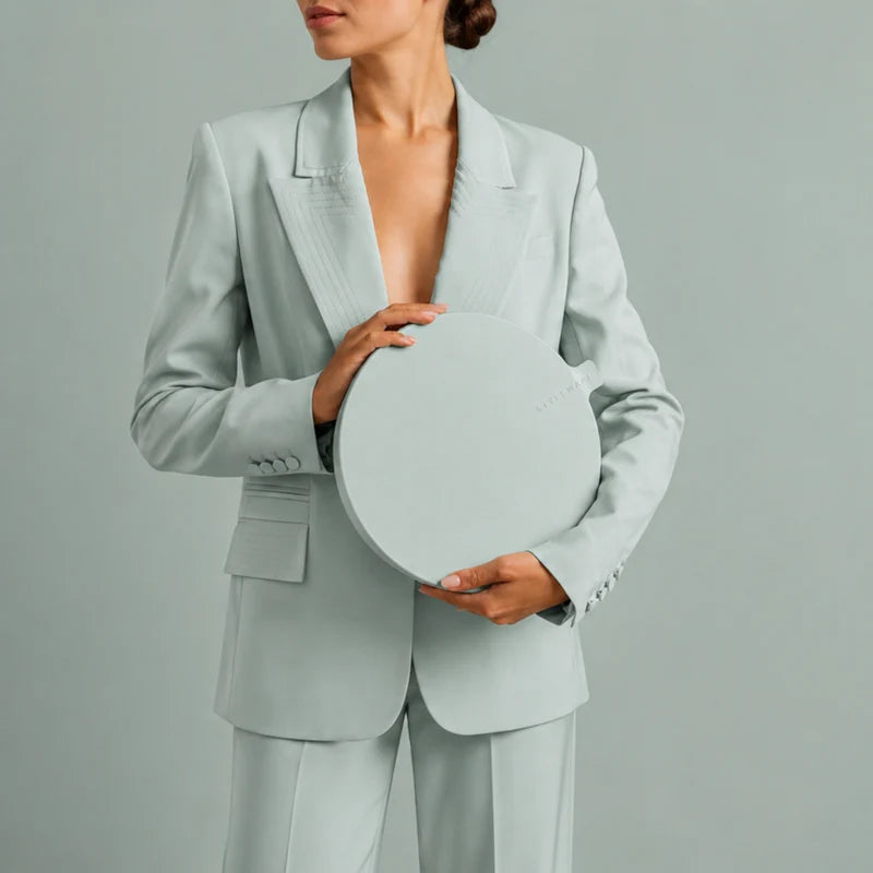 A model wearing a light sage green tailored blazer and matching trousers, holding a matching round sage green container against a solid light sage background.
