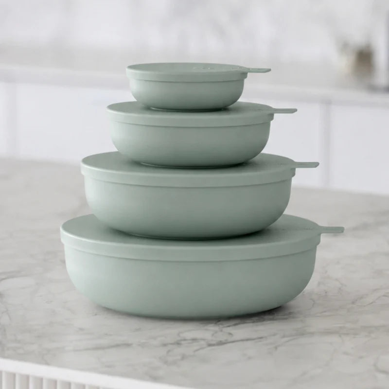 Four nested round storage containers with matching lids in a muted sage green color, arranged on a light marble countertop with a softly blurred kitchen background.