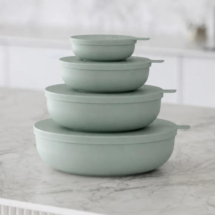 Four nested round storage containers with matching lids in a muted sage green color, arranged on a light marble countertop with a softly blurred kitchen background.