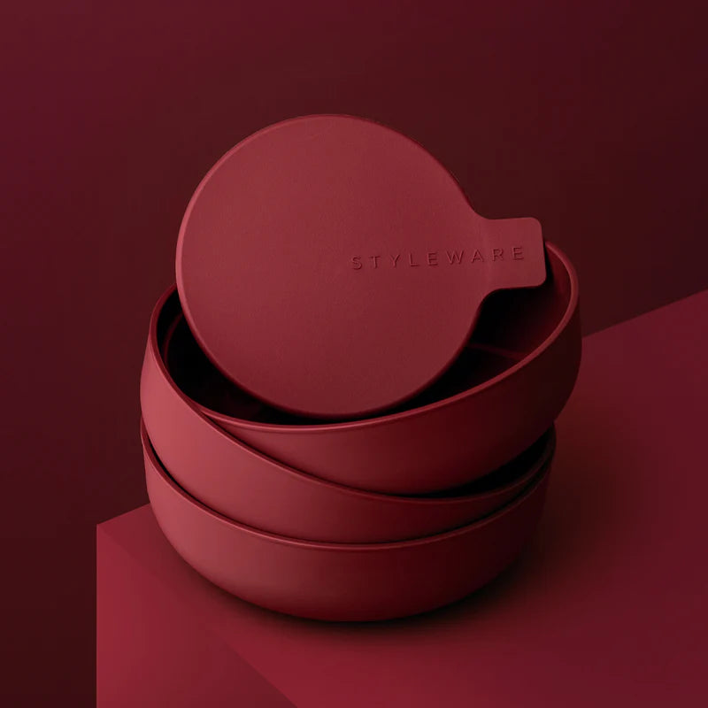 A stack of matte red food storage bowls with a matching lid resting on top, featuring the embossed brand name 'STYLEWARE' on the lid, set against a matching dark red background.