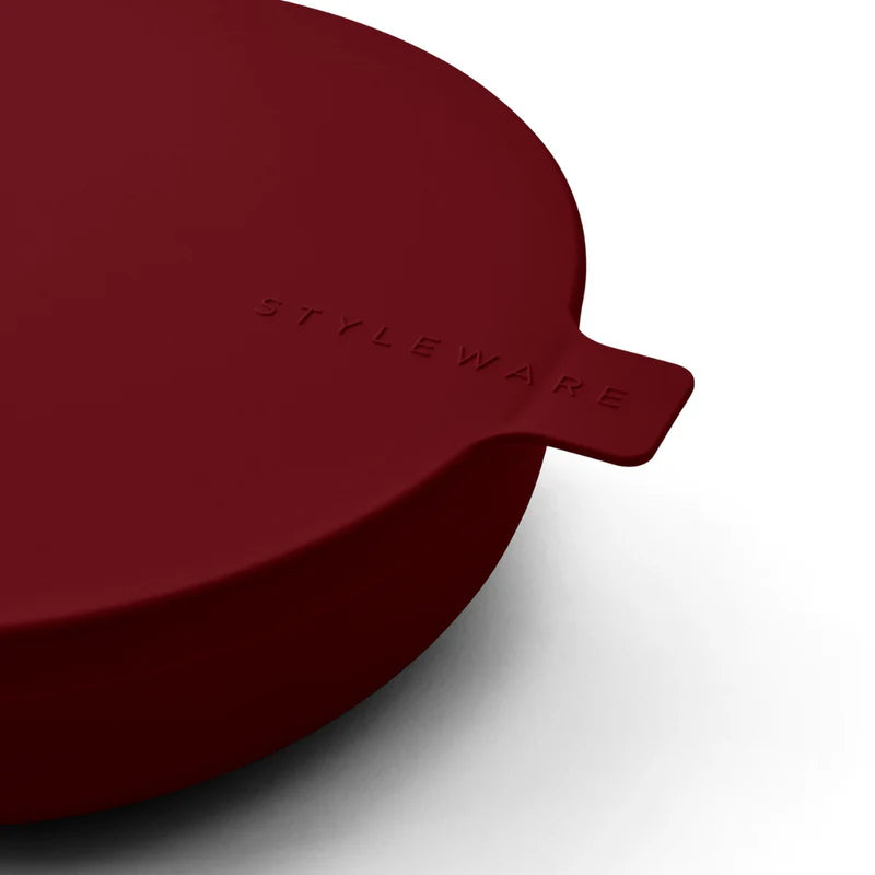 A close-up of a dark red bowl lid with a small tab handle, embossed with the brand name &