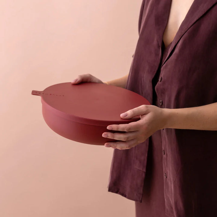A person in a dark plum button-front top holds a matte terracotta red oval container with a matching lid and a small side tab, against a soft peach-pink background.
