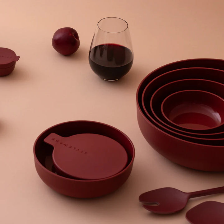A coordinated set of matte deep red dinnerware and tableware arranged on a warm beige surface, including nested bowls, a lidded bowl with a spoon inside, a wine glass with red wine, and a spoon. No person or real environment is present, so this is Product/Packaging rather than Lifestyle.