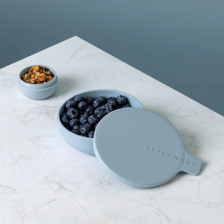 A light blue container with a matching lid, shown open with blueberries inside on a white marble surface. A small matching container with granola sits in the background.