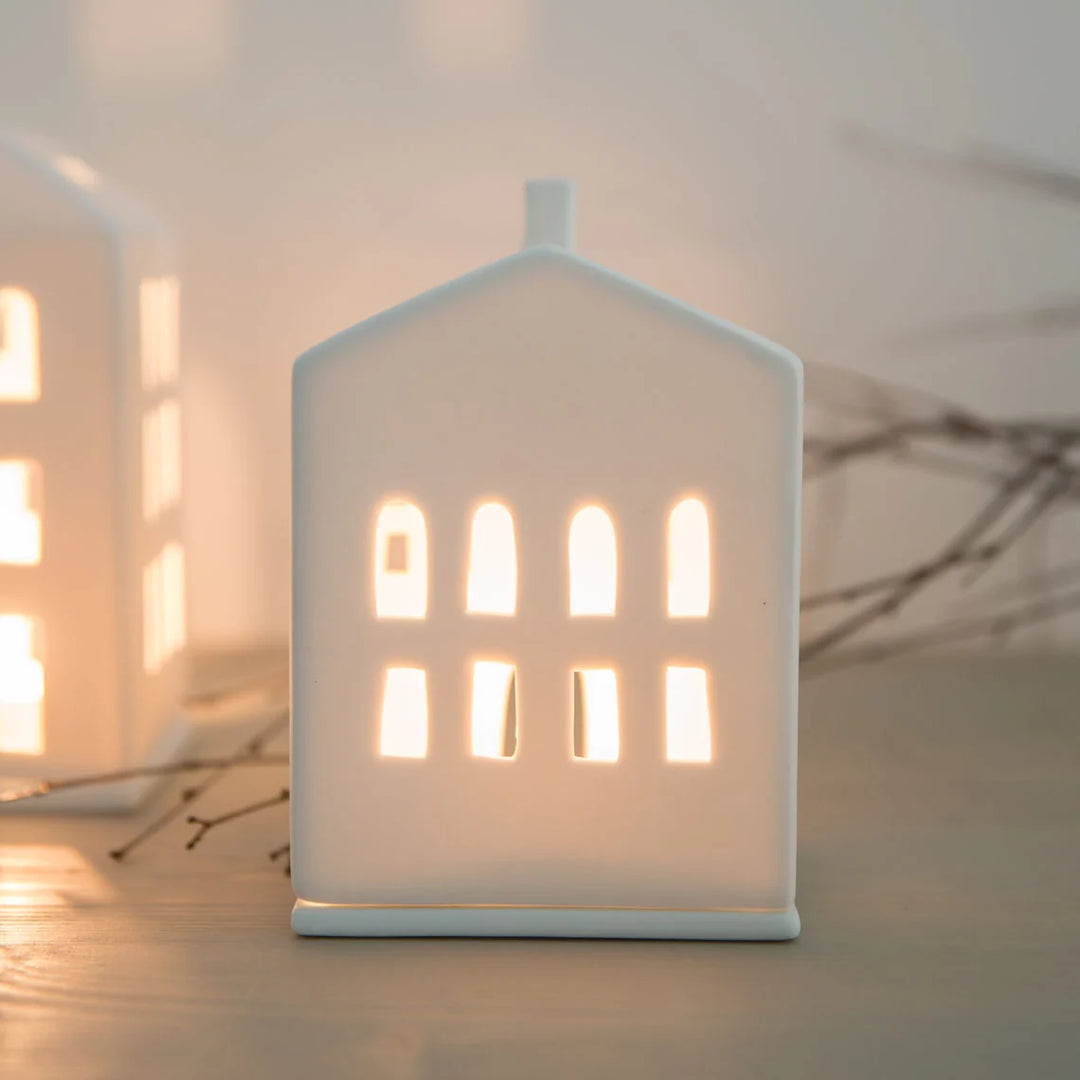 A small white house-shaped ceramic tea light holder with arched window cutouts, glowing warm from an internal light, set against a softly blurred neutral background.