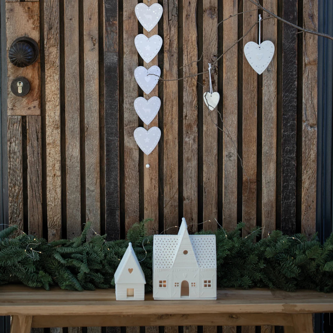 Two white gingerbread house shaped tealight candle holders on a wood bench in front of a timber paling fence