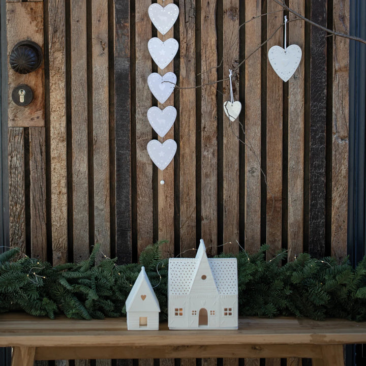 Two white gingerbread house shaped tealight candle holders on a wood bench in front of a timber paling fence